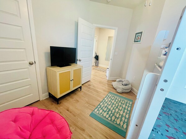 A room with a television on a cabinet, a pink bean bag, a green mat with fruit patterns, and a white door.