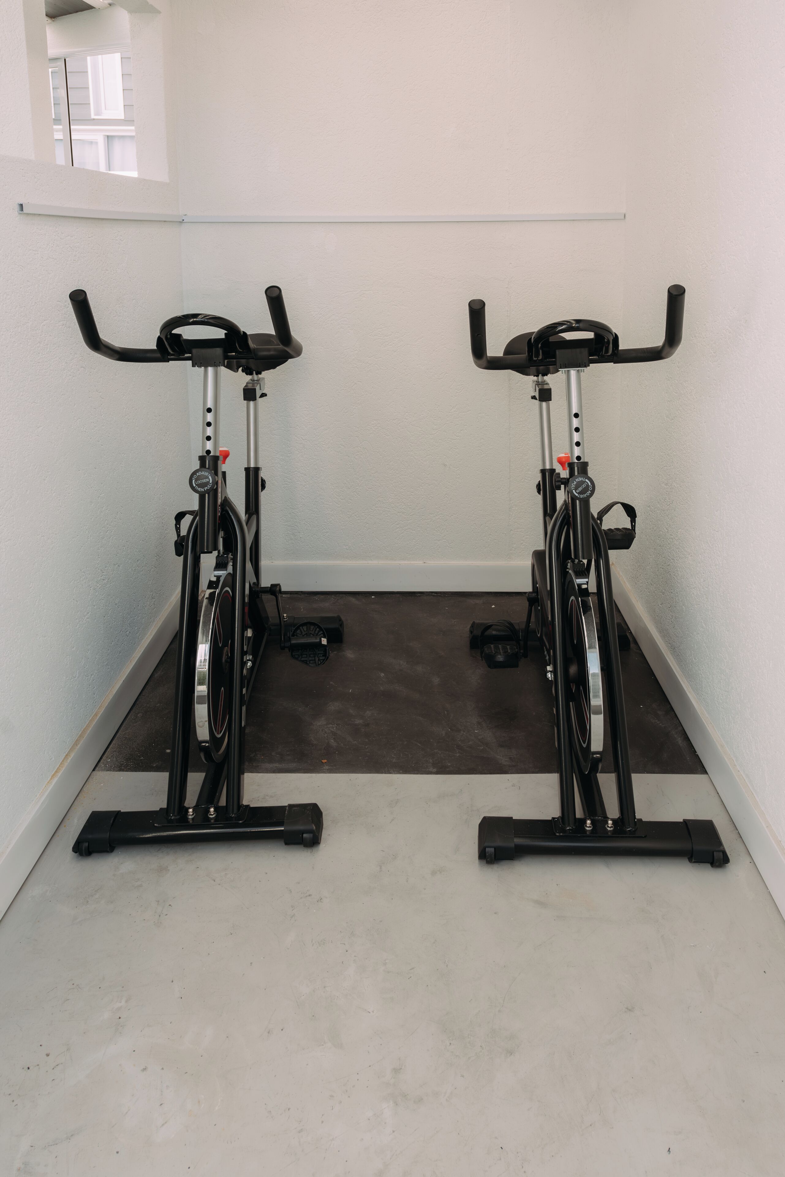 Deux vélos d’appartement sont placés dans un couloir étroit, avec un mur blanc et une fenêtre au fond.
