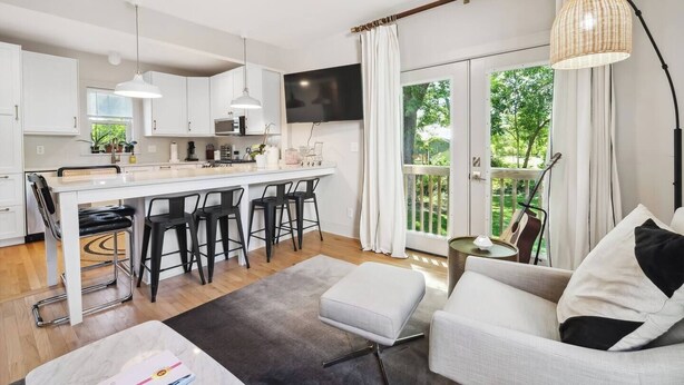 A modern kitchen with white cabinets, a central island with bar stools, a flat-screen TV, and a balcony with a view of greenery. - Montrose Manor- In the heart of Belmont! (Charlottesville)
