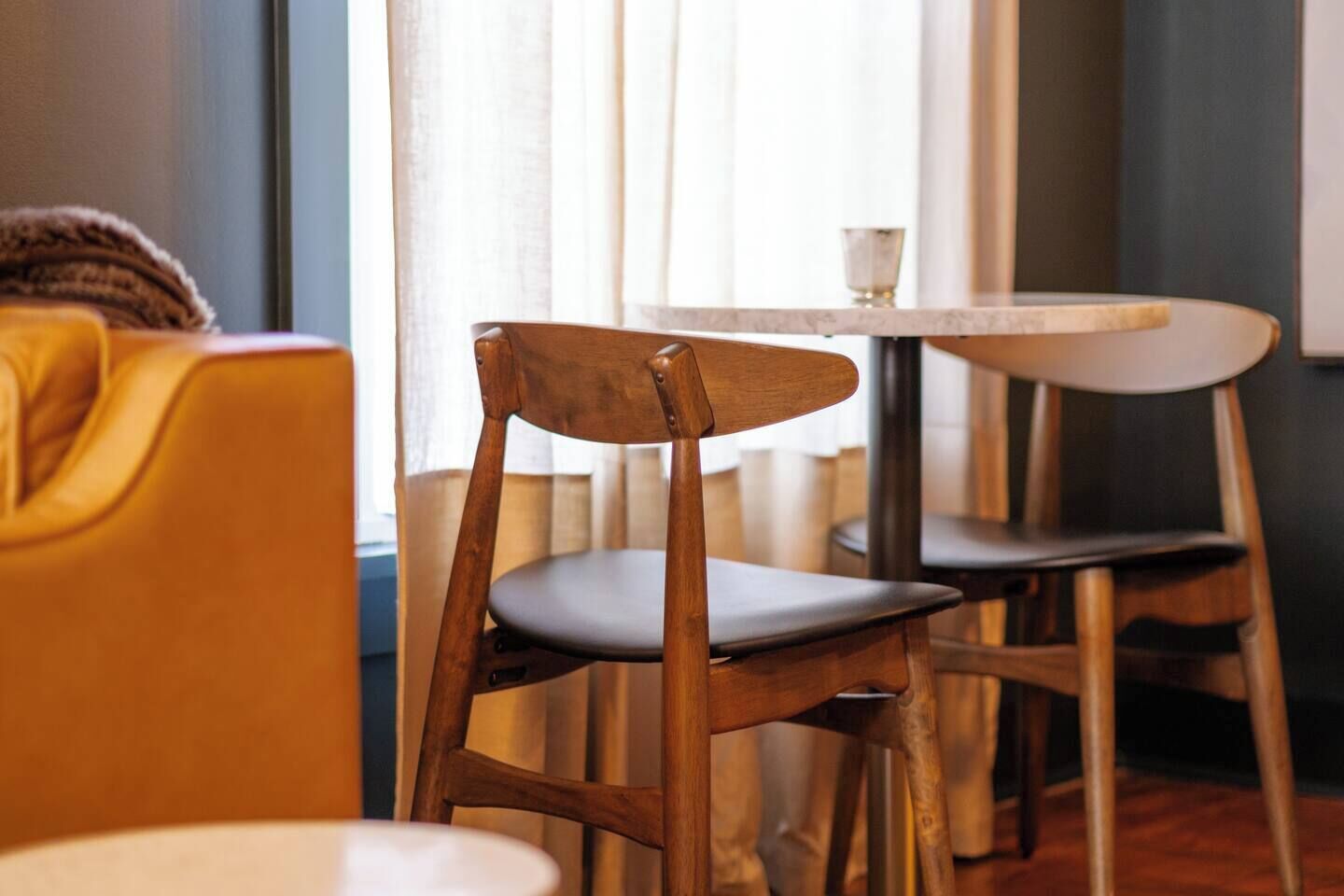 Une chaise en bois avec un siège foncé, une table ronde blanche et un canapé jaune dans une pièce dotée d’une fenêtre et de rideaux.