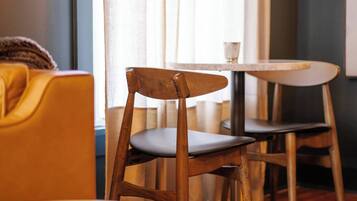 Une chaise en bois avec un siège foncé, une table ronde blanche et un canapé jaune dans une pièce dotée d’une fenêtre et de rideaux.