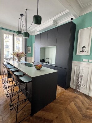Uma cozinha moderna com uma ilha central, banquetas, luminárias pendentes e vista da cidade por grandes janelas.