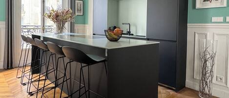 Een moderne keuken met een centraal eiland, barstoelen, hanglampen en een stadszicht door grote ramen.