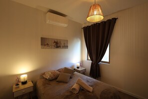A bedroom with a bed, a bedside table with a lamp, a window with curtains, and a wall-mounted air conditioner.