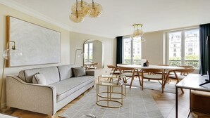 A modern living room with a sofa, dining table, and chairs. There is a large window with a view of buildings.