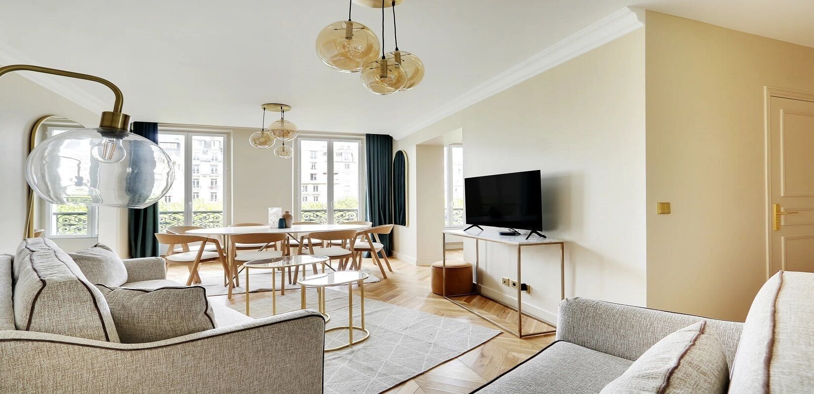 Un salon moderne comprenant un canapé, une table à manger avec des chaises, un téléviseur posé sur un meuble et une grande fenêtre avec des rideaux.