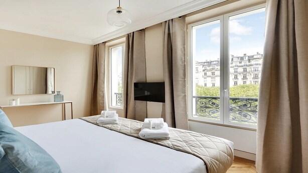 A hotel room with a large bed, a TV mounted on the wall, a window with a view of buildings, and a balcony with a railing.