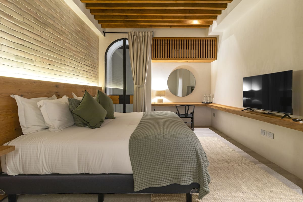 A bedroom with a wooden headboard, a bed with white and green bedding, a television on a wooden shelf, a round mirror, and a window with curtains.