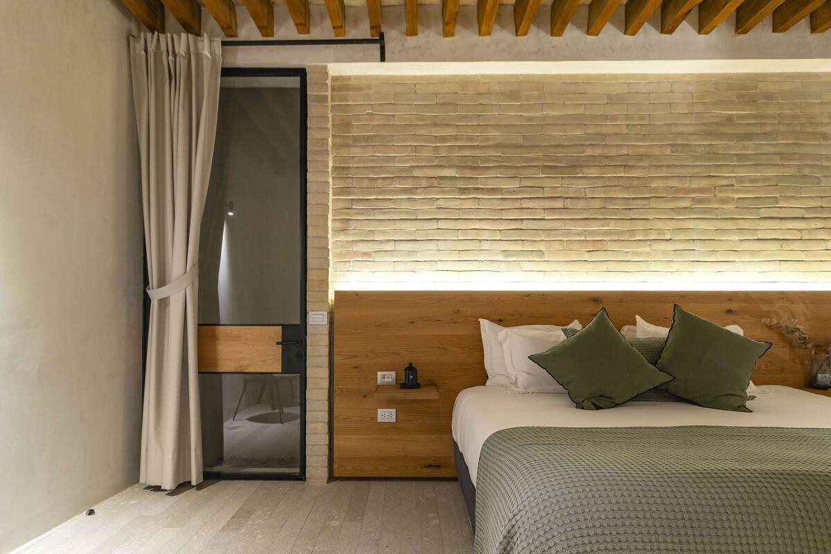A modern bedroom with a wooden headboard, a bed with green pillows, a nightstand, and a glass door leading to a bathroom.
