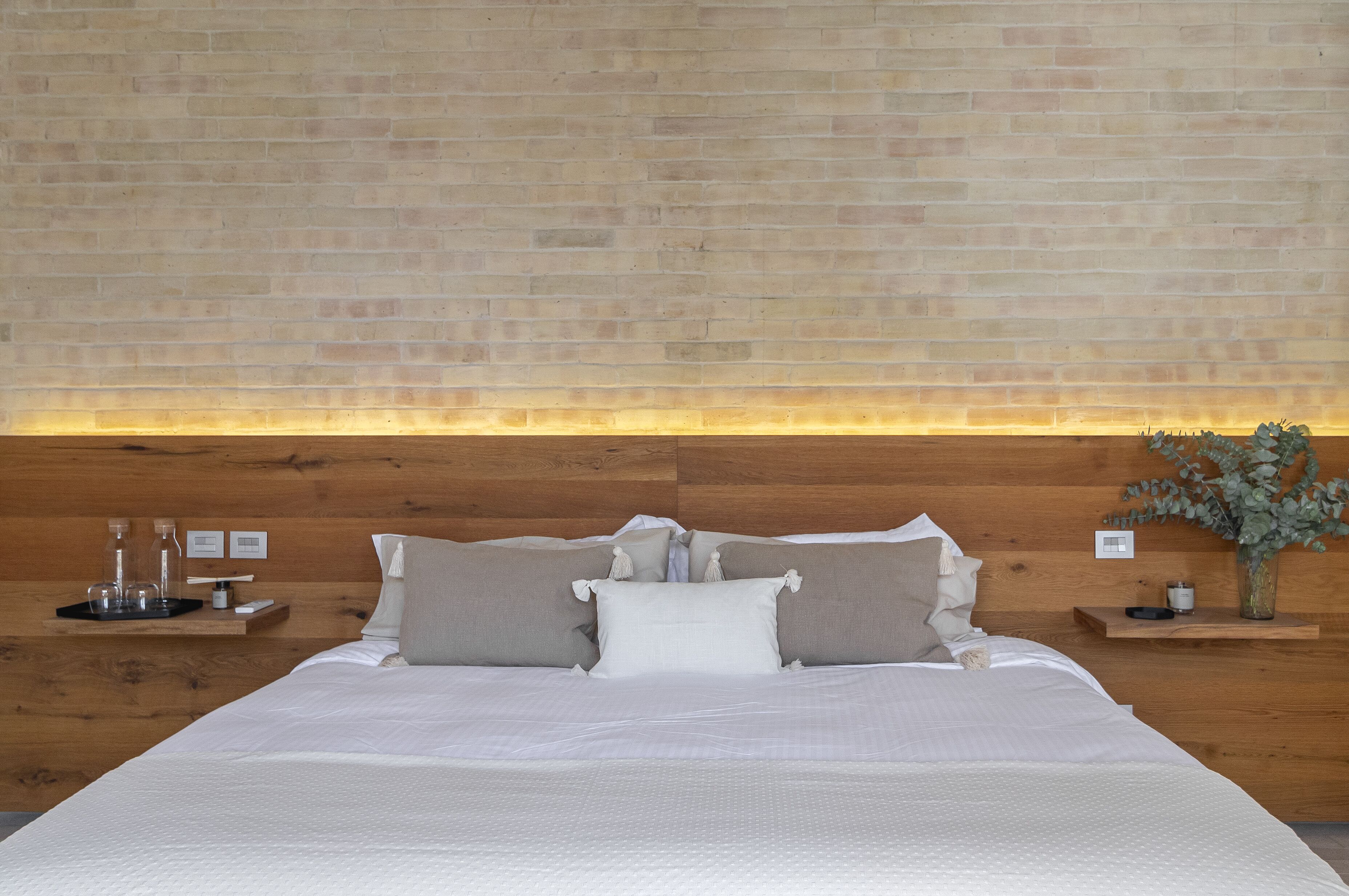 Une chambre à coucher bien aménagée, avec une tête de lit en bois, un grand lit recouvert de linge de lit blanc, deux tables de chevet ornées d’objets décoratifs et un luminaire fixé au mur.