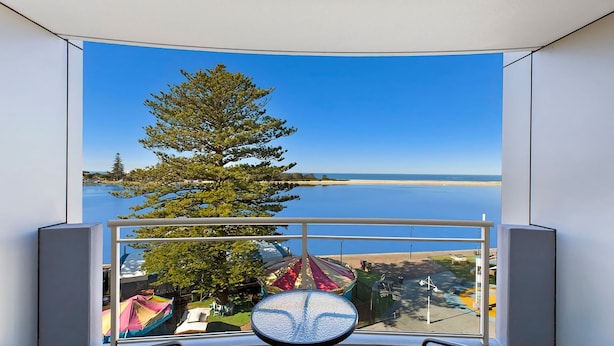 A room with a curved window offering a view of a calm body of water, a large tree, and a distant shoreline. - The Entrance Waterfont Apartment 303 (The Entrance)