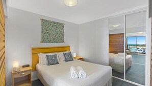 A hotel room with a bed, bedside tables, a mirror, and a view of the beach. - The Entrance Waterfont Apartment 303 (The Entrance)