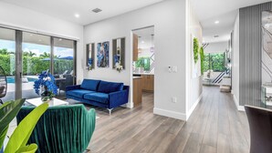 Un espace de vie moderne avec un canapé bleu, une table à manger recouverte d’une nappe verte, et une vue sur une piscine et la verdure environnante.