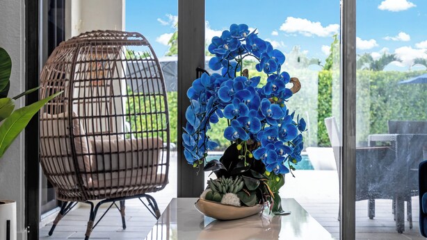 Un intérieur moderne avec une porte vitrée donnant sur un balcon, une chaise en osier et un vase de fleurs bleues posé sur une table.