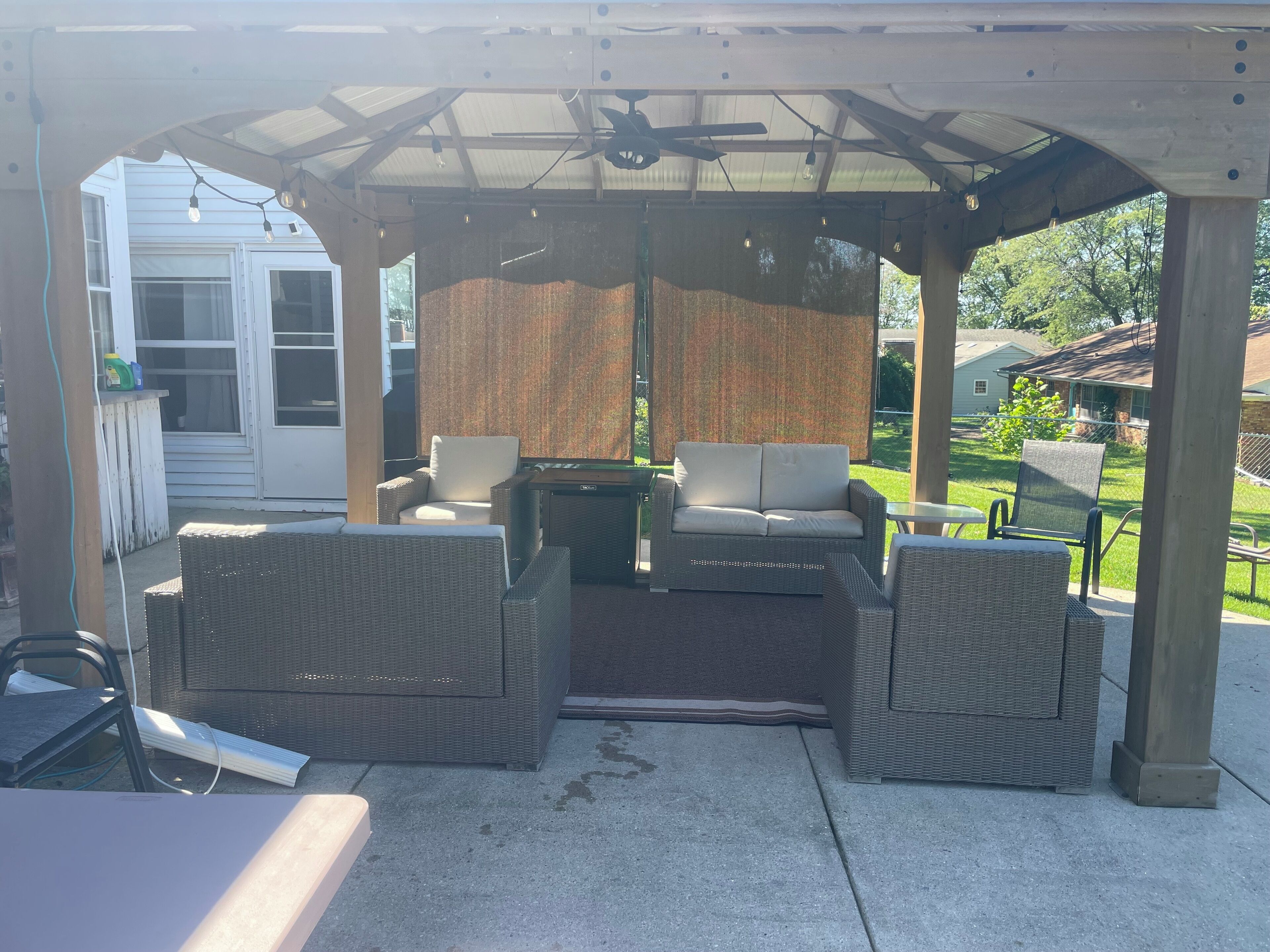 A covered patio area with wicker furniture, including a sofa and chairs, under a pergola with a ceiling fan.