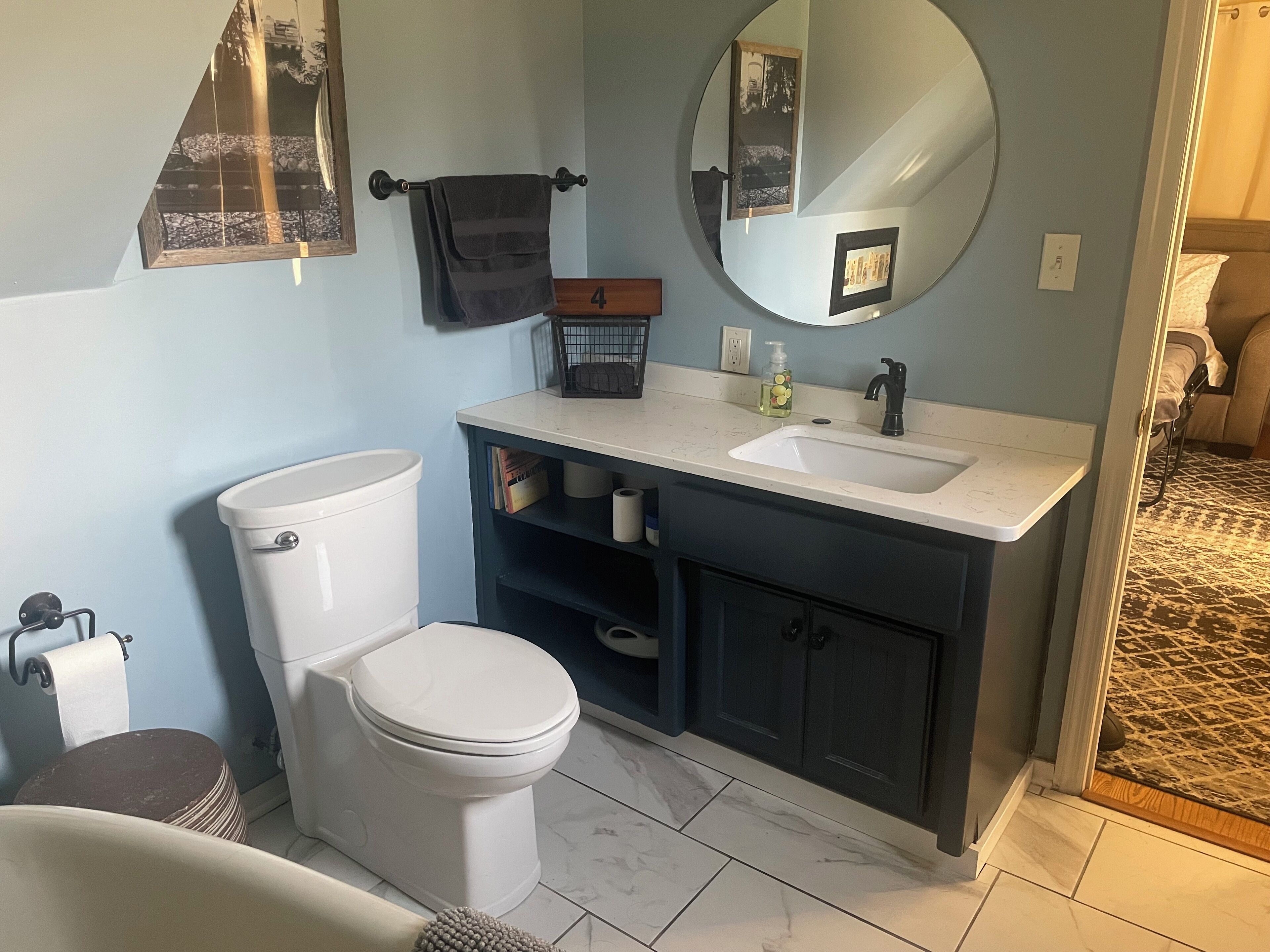 A bathroom with a toilet, sink, and mirror. There is a towel rack with a towel, a framed picture on the wall, and a small shelf with toiletries.