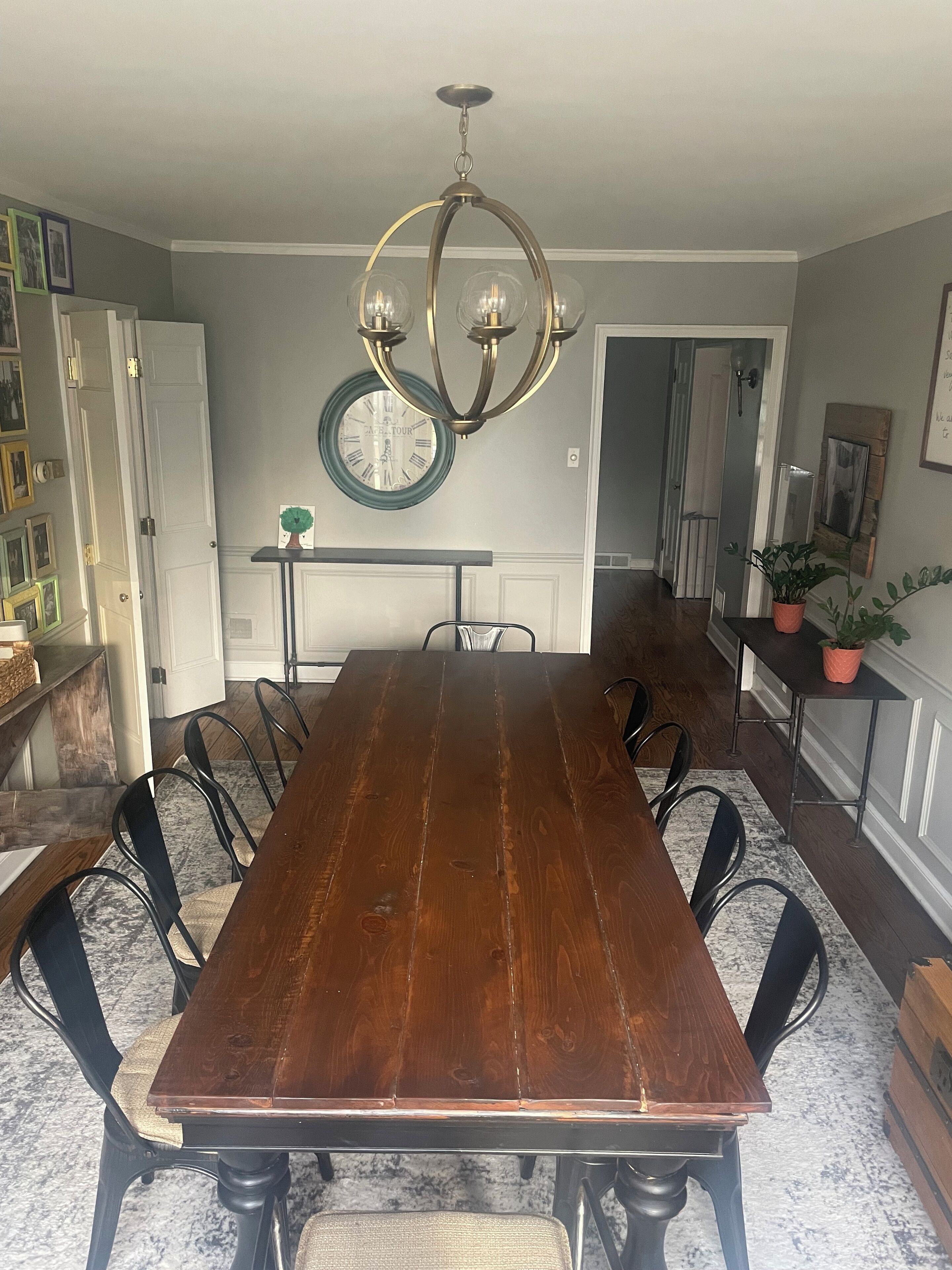 Une salle à manger avec une longue table en bois, des chaises noires, un lustre, une horloge et une plante en pot.