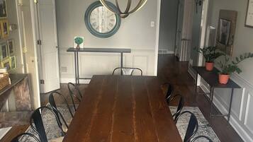 Une salle à manger avec une longue table en bois, des chaises noires, un lustre, une horloge et une plante en pot.