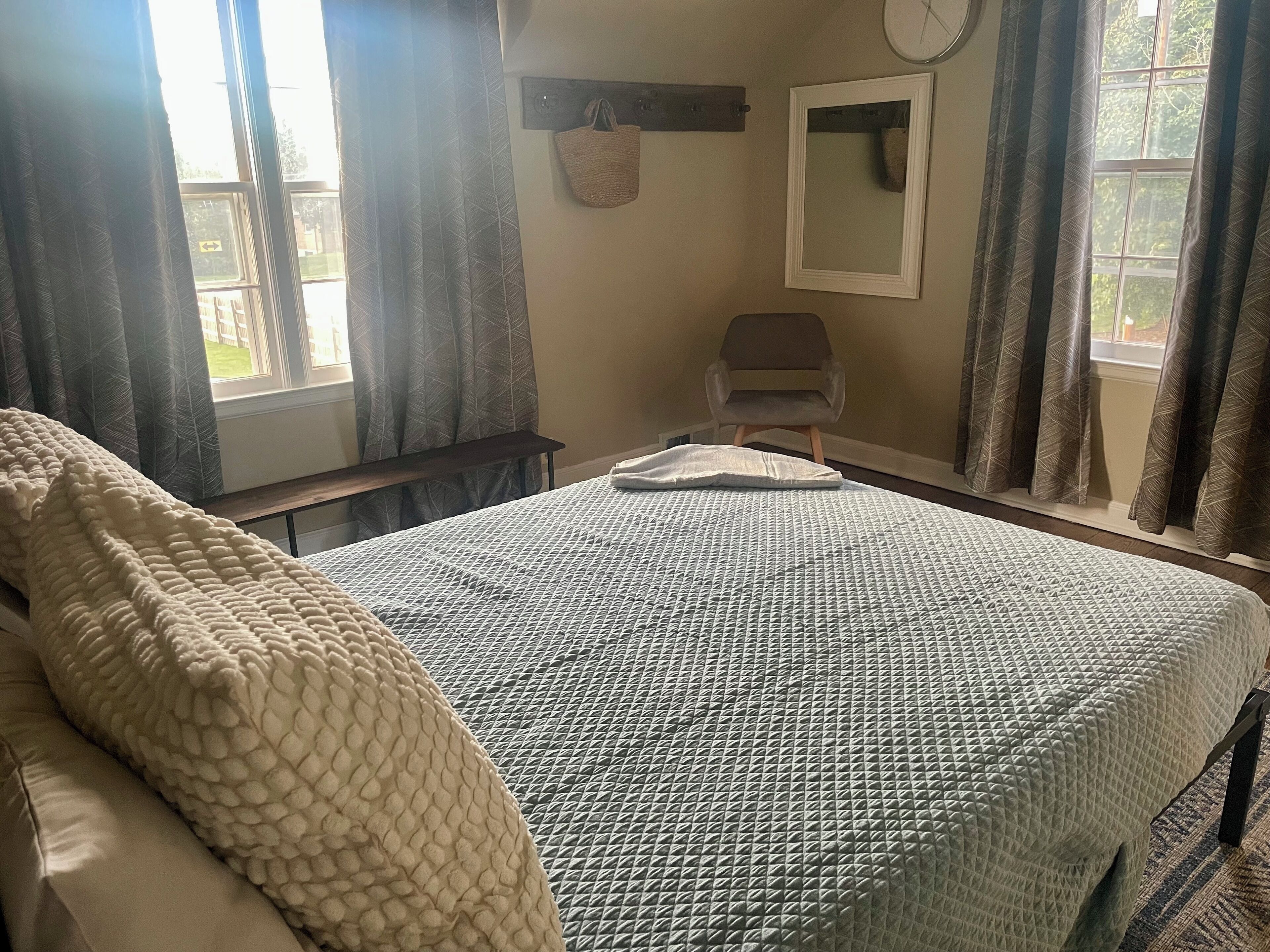 A neatly made bed with a patterned bedspread, a chair, a wall-mounted mirror, and a window with curtains.