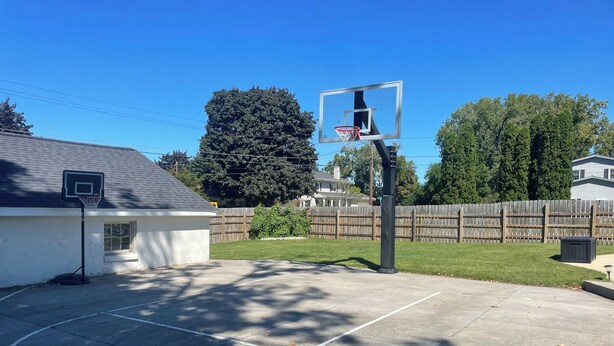 Una cancha de baloncesto con aro, rodeada por una cerca de madera, un cobertizo pequeño y un árbol grande.