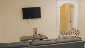 En stue med en grå sofa, et fladskærms-tv ophængt på væggen, et lille bord med en højttaler og fjernbetjening, samt en lænestol med et mønster.