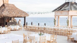 Un salón de bodas al aire libre con mesas y sillas redondas, una estructura de techo de paja y un pabellón con techo abovedado.