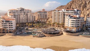 Un hotel frente al mar con varias construcciones, una playa de arena y un acantilado rocoso al fondo.