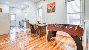 A spacious room with a wooden floor, a foosball table, a dining area with chairs and a table, a kitchen in the background, and a ceiling fan. - Stylish 2BR Minutes to Downtown - Prime Location (Savannah)