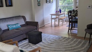 A spacious living area with a sofa, a wooden chair, a dining table, and a large window offering a view of greenery.