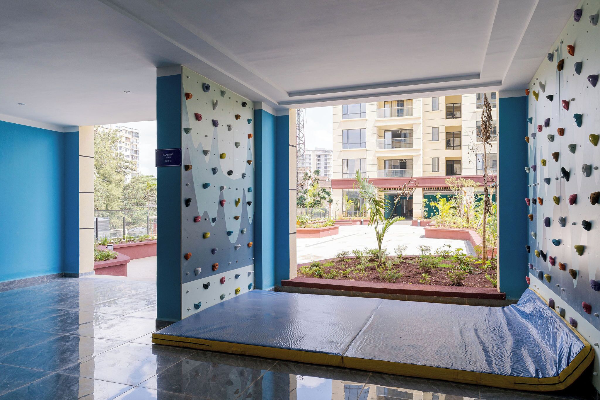 A climbing gym with a padded floor and climbing walls, a blue wall, and a view of a building and greenery outside.