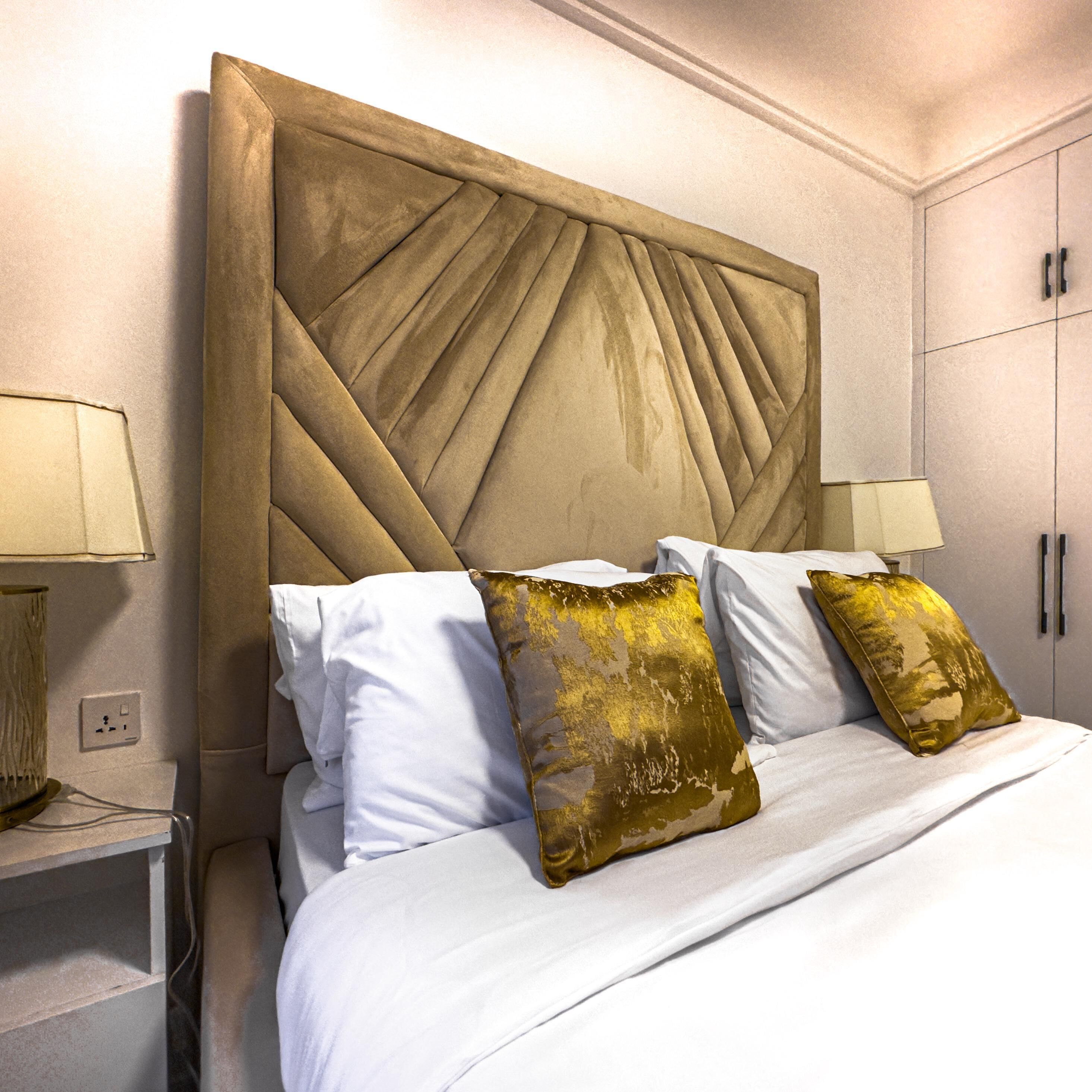 Una cama bien hecha con sábanas blancas y almohadas decorativas, un amplio cabecero tapizado y una mesita de noche con lámpara.