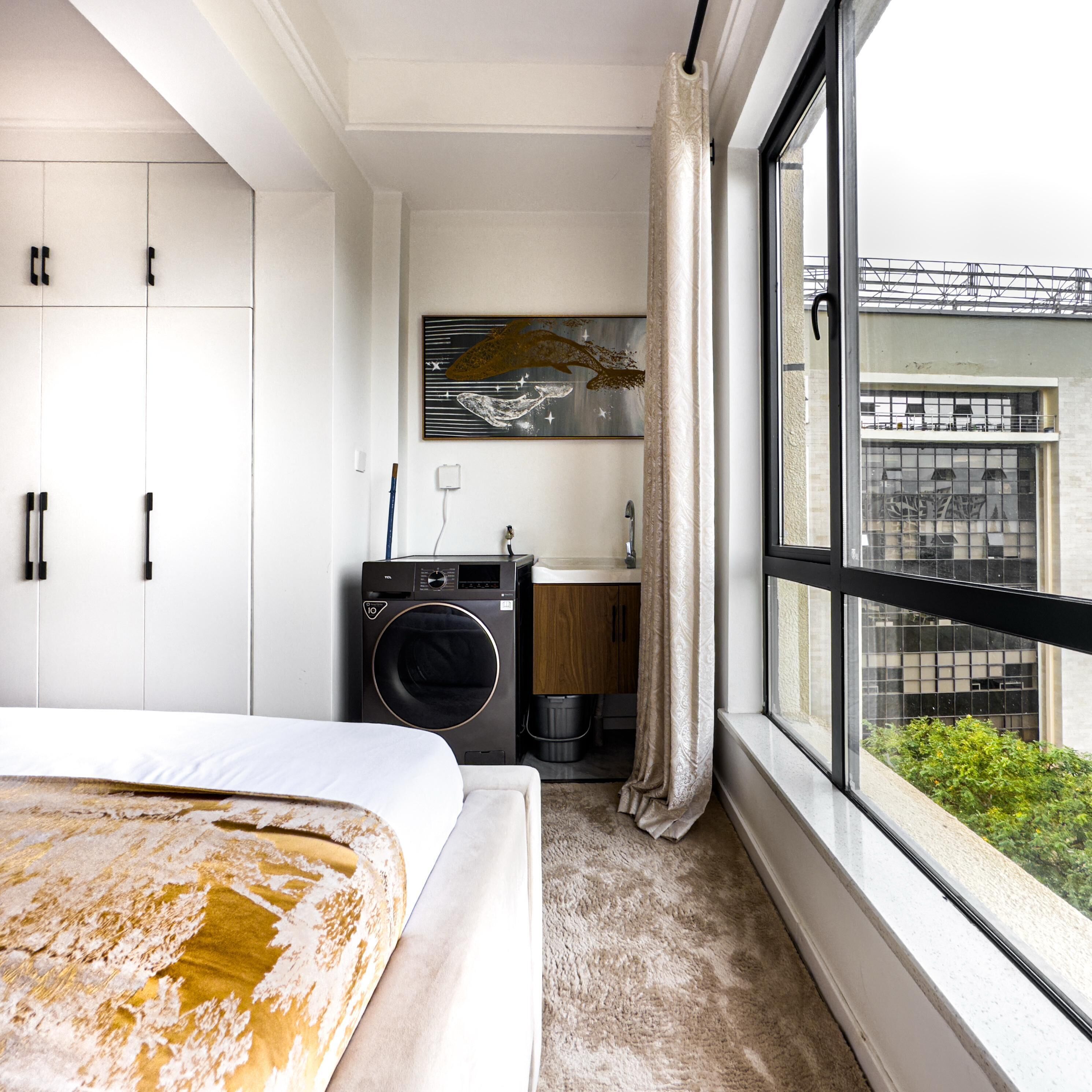 A hotel room with a bed, a washing machine, a sink, and a large window with a view of buildings.