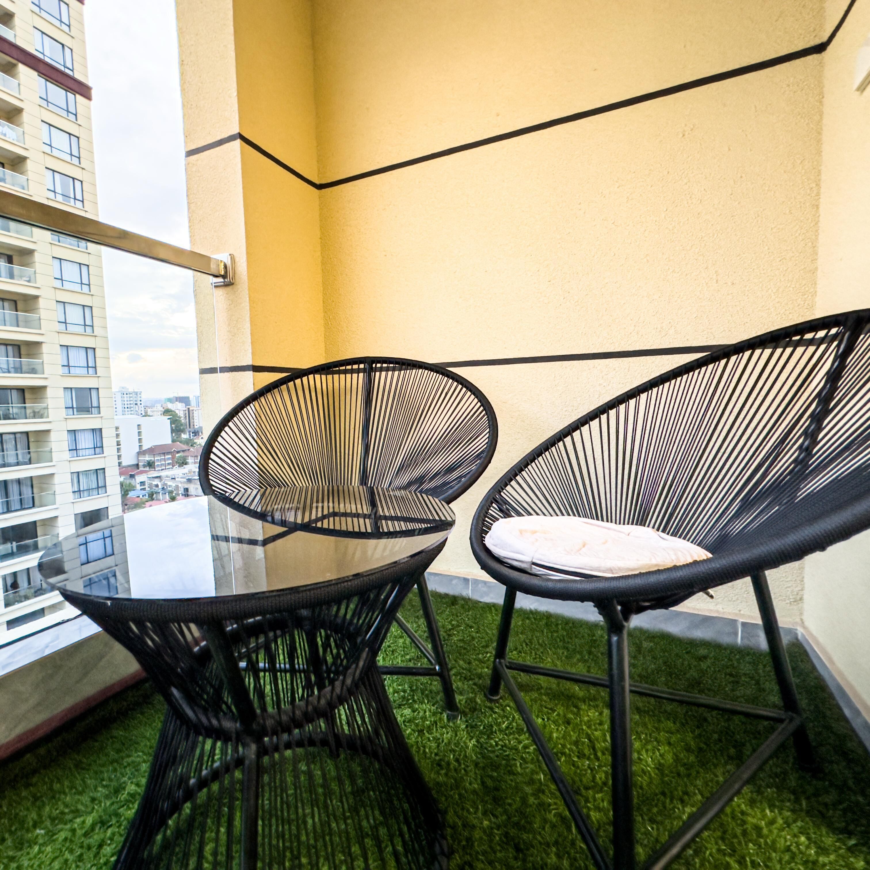 Un balcon avec deux chaises en métal noir à mailles et une table en verre, offrant une vue sur le paysage urbain à travers une porte vitrée.