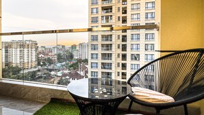 Un balcón con una mesa de vidrio y dos sillas negras, con vistas a un paisaje urbano de edificios altos.