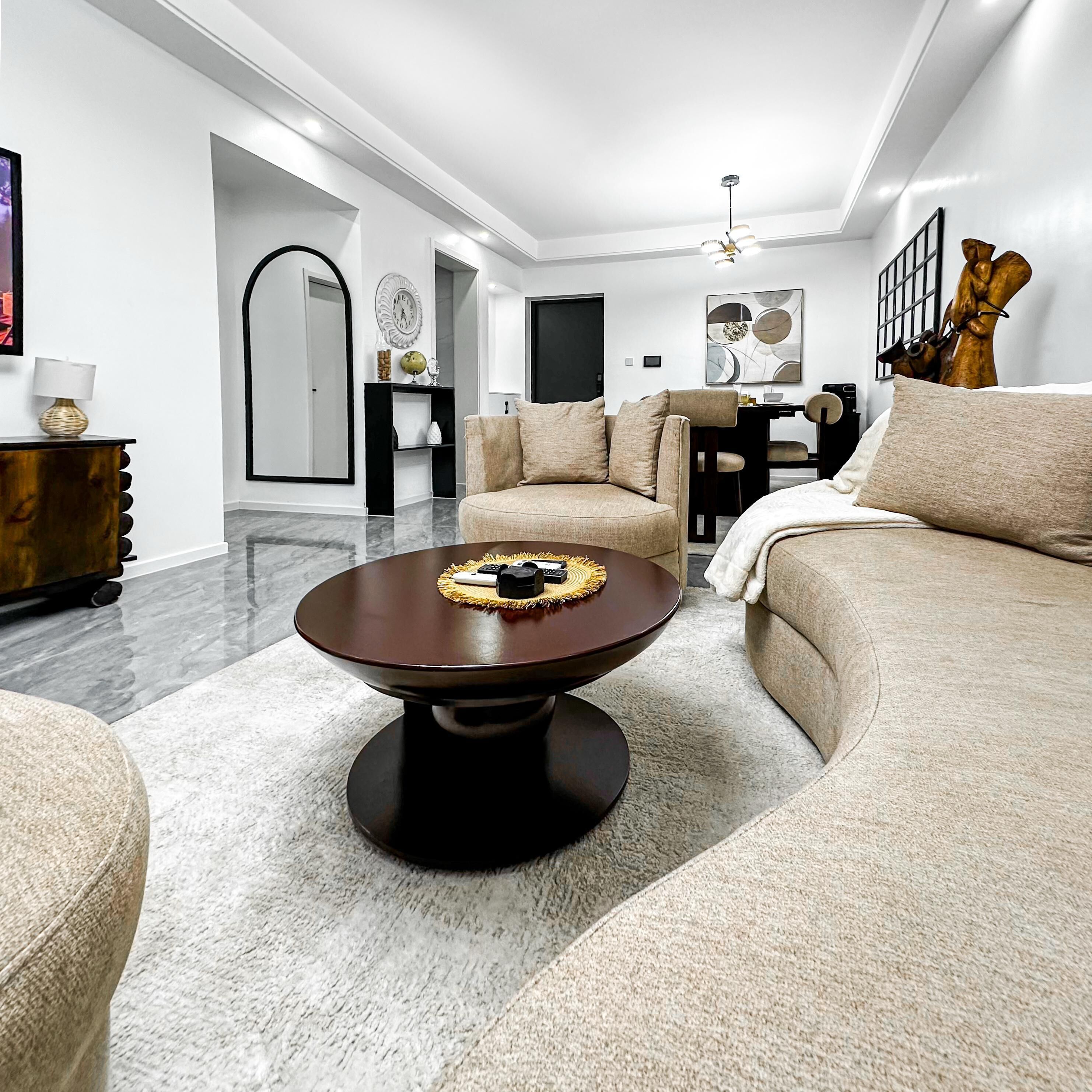 A modern living room with a round coffee table, beige sofas, a dining area with a black table, and a large mirror.