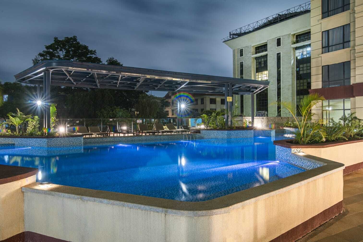 A swimming pool with a covered area and lounge chairs, surrounded by a building with multiple floors and windows.
