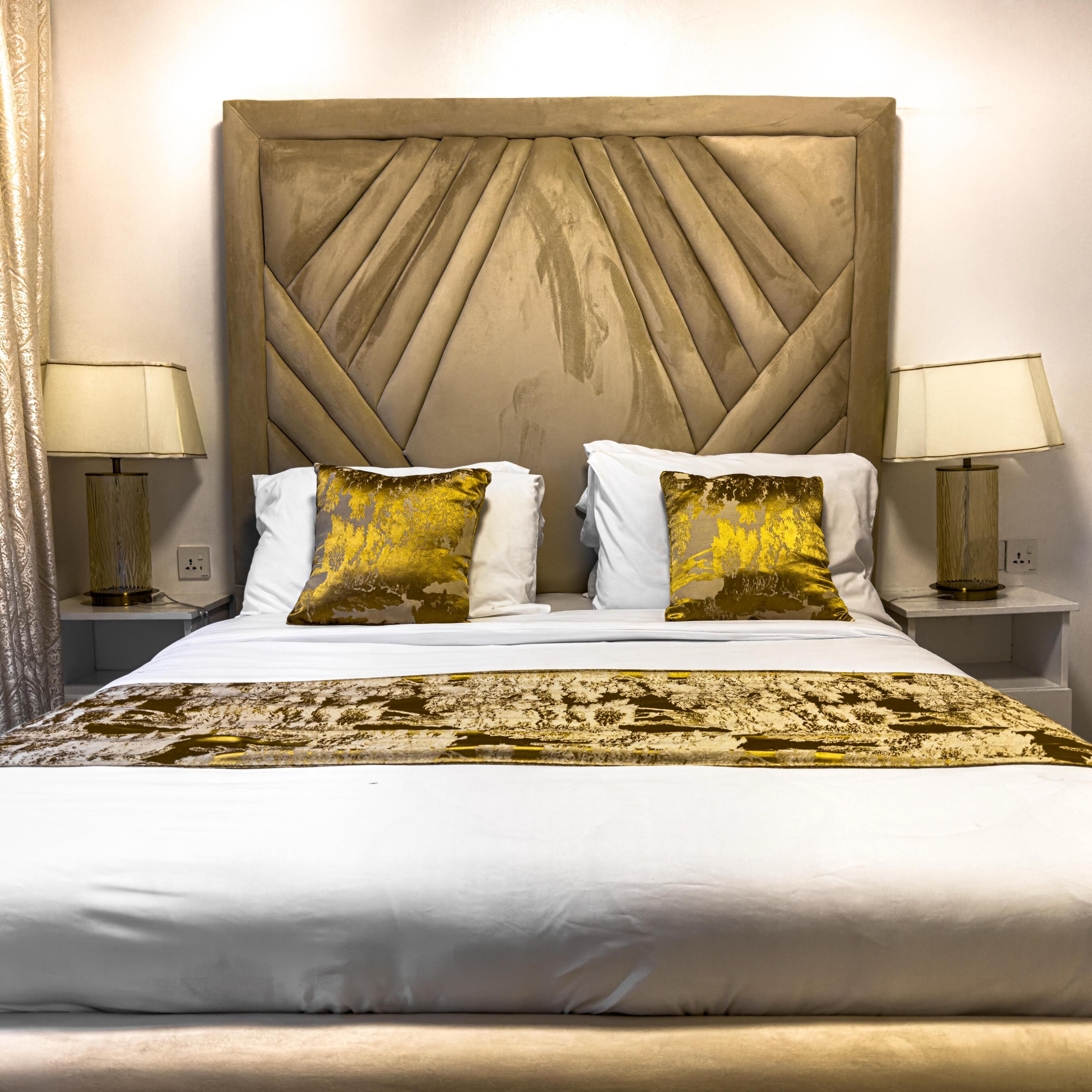 Een net opgemaakt bed met witte lakens, twee kussens en een plaid in goud- en witmotief. Aan beide zijden staan nachtlampjes met beige kapjes.