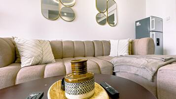 A living room with a sectional sofa, a coffee table with a decorative vase and remote controls, and a refrigerator in the background.