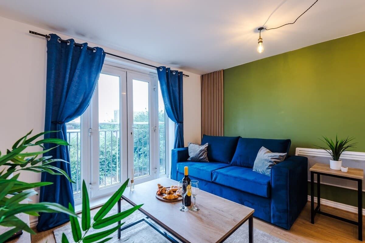 A living room with a blue sofa, a coffee table with a bottle and glasses, and a view through a window with curtains.