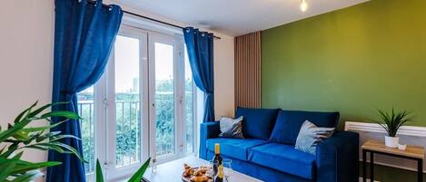 A living room with a blue sofa, a coffee table with a bottle and glasses, and a view through a window with curtains.