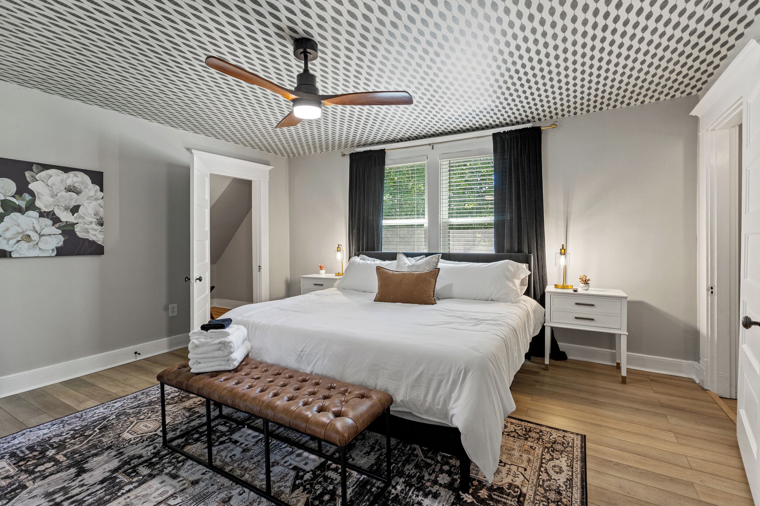 A bedroom with a bed, a bench, a ceiling fan, a nightstand, and a window with curtains.