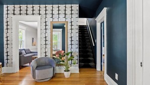 A living room with a patterned wallpaper, a large mirror, a grey armchair, and a potted plant.