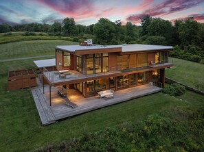 Une maison moderne à deux étages, dotée d’une terrasse, de grandes fenêtres et offrant une vue sur le paysage environnant.