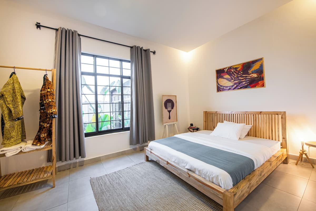 A bedroom with a large bed, a window with curtains, a wooden headboard, a painting on the wall, and a clothing rack.