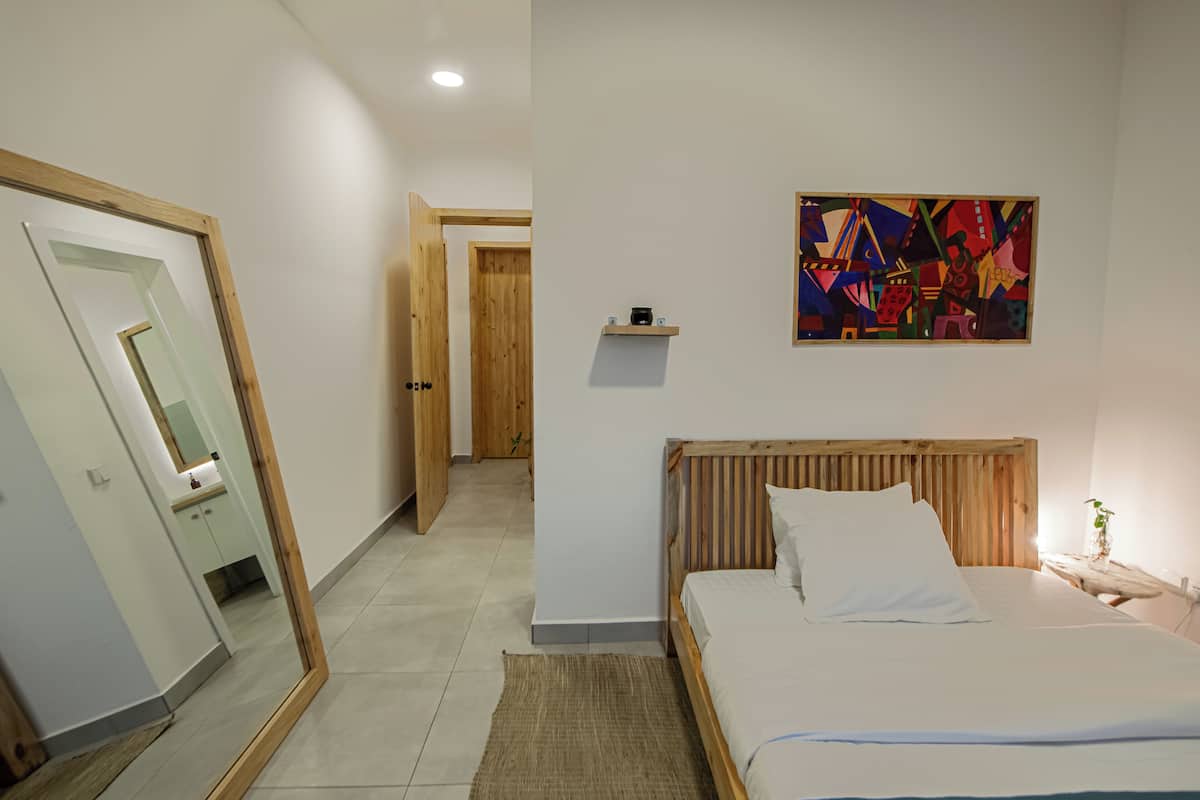 A bedroom with a wooden headboard, a bed with white linens, a small bedside table with a lamp, a mirror on the wall, and a colorful abstract painting.