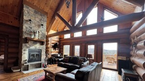 A log cabin interior with a stone fireplace, leather sofas, and a large window overlooking a deck.