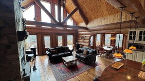 A spacious log cabin interior with a large living area, wooden floors, and a dining area with a view of the outdoors.