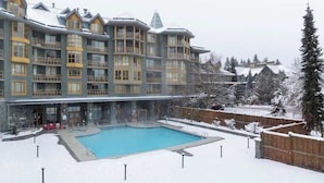 A multi-story hotel with a pool area, surrounded by snow-covered trees and a wooden fence.