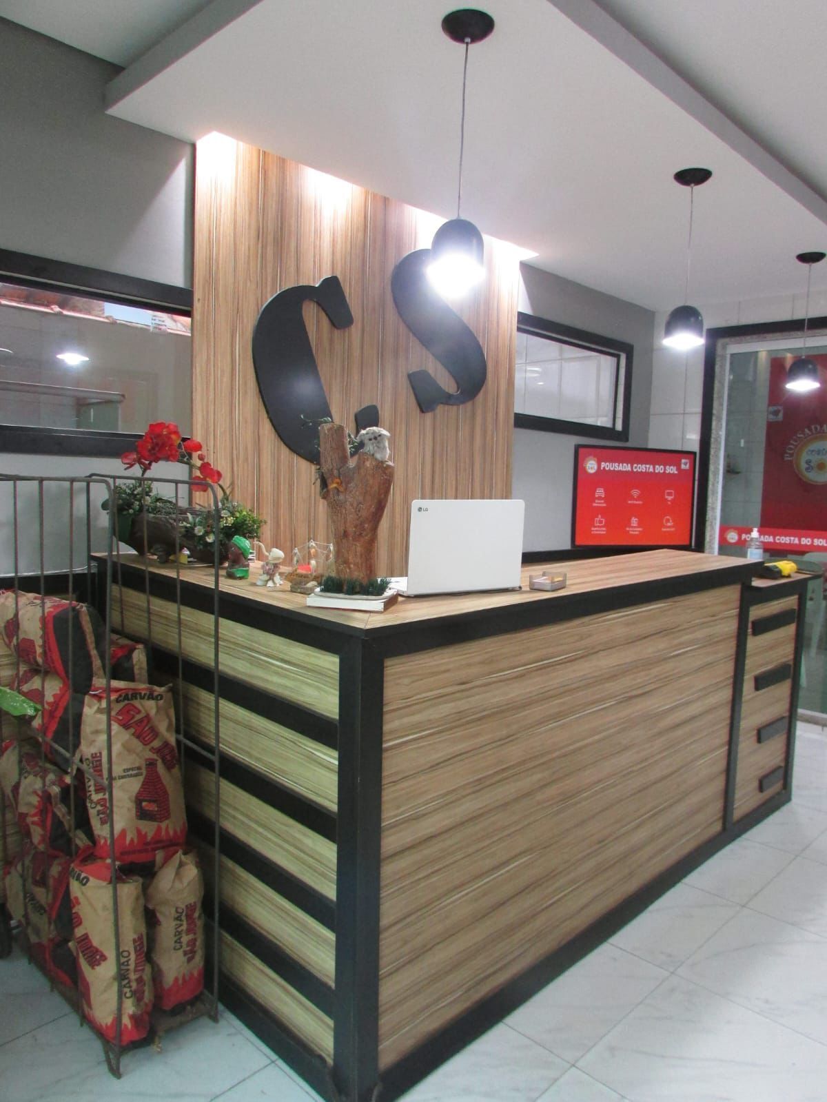 Una recepción con detalles de madera, un gran logotipo en forma de 'S' y una pantalla digital que muestra 'Pousada Costa do Sol'.
