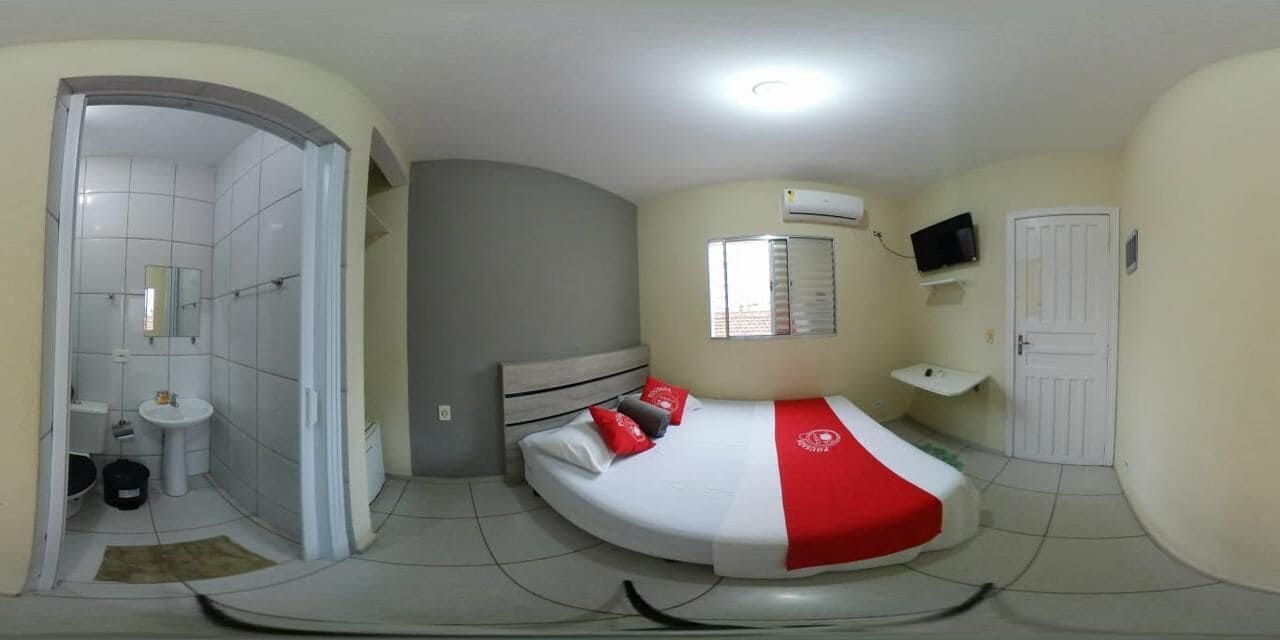 A compact bedroom with a curved bed, a small bedside table, a window with blinds, and a TV mounted on the wall.